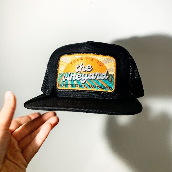 An image of a flat brim hat from Brasswood Estate, with an embroidered patch of a sunrise over a vineyard, with text that reads, "Take Me To The Vineyard - Brasswood Estate, Napa Valley, CA.