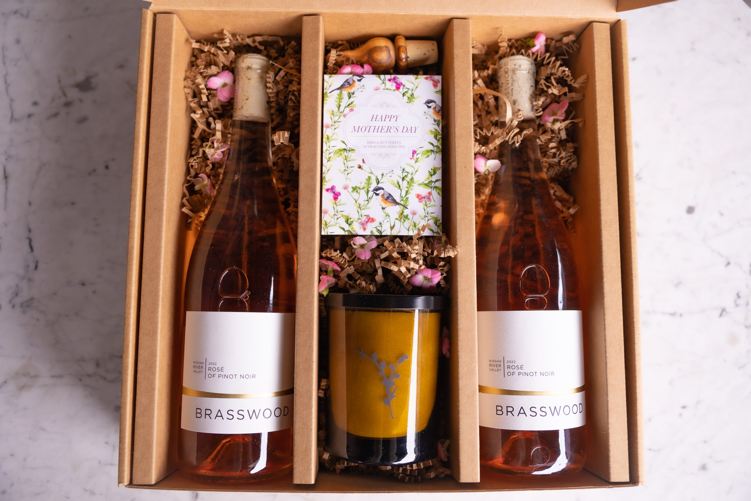 Photo of a gift box with two Brasswood Cellars' Rosé of Pinot Noir bottles, a candle, and wildflower seeds packet.