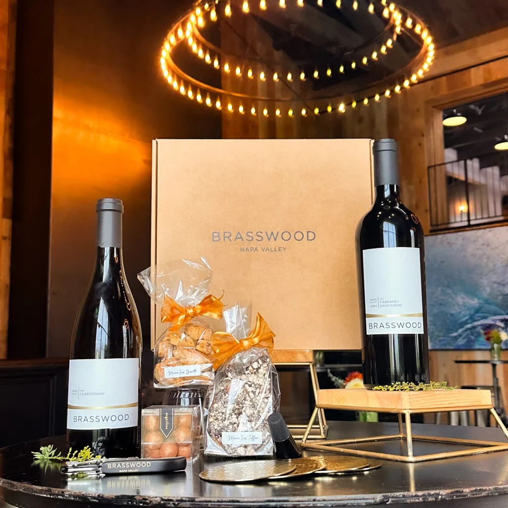 A photo of the Brasswood 2024 Winter Box which features the 2022 Chardonnay, the 2021 Cabernet Sauvignon, and various candies and treats, and neatly placed on a circular table in the Brasswood Event Dining Space.